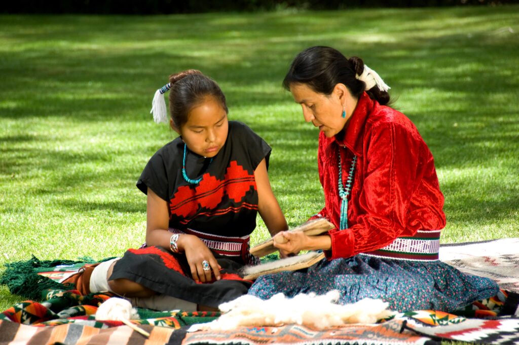Indigenous elder teaching a young girl traditional weaving outdoors, illustrating intergenerational learning and cultural resilience