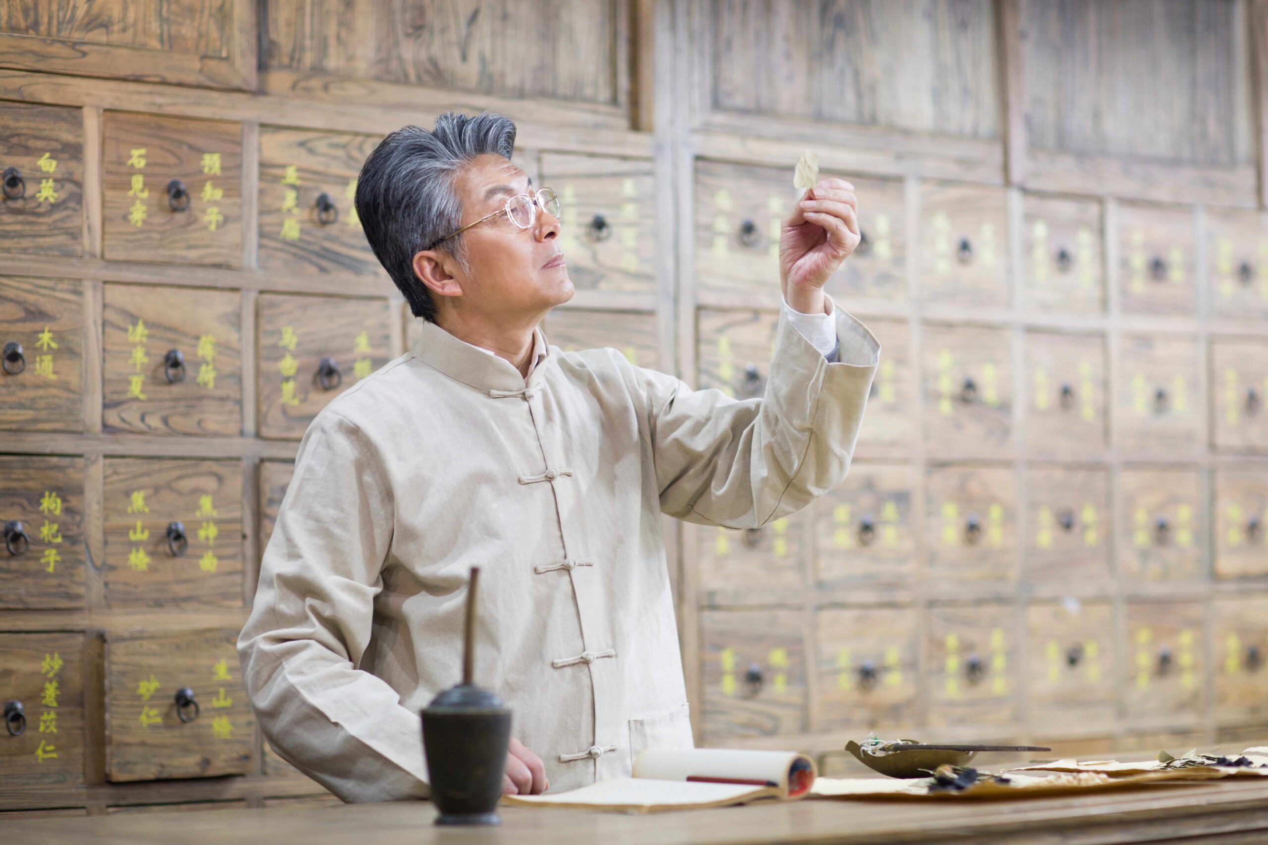 A traditional Chinese medicine practitioner examines a dried medicinal herb in a wooden apothecary with labeled drawers used for herbal medicine.