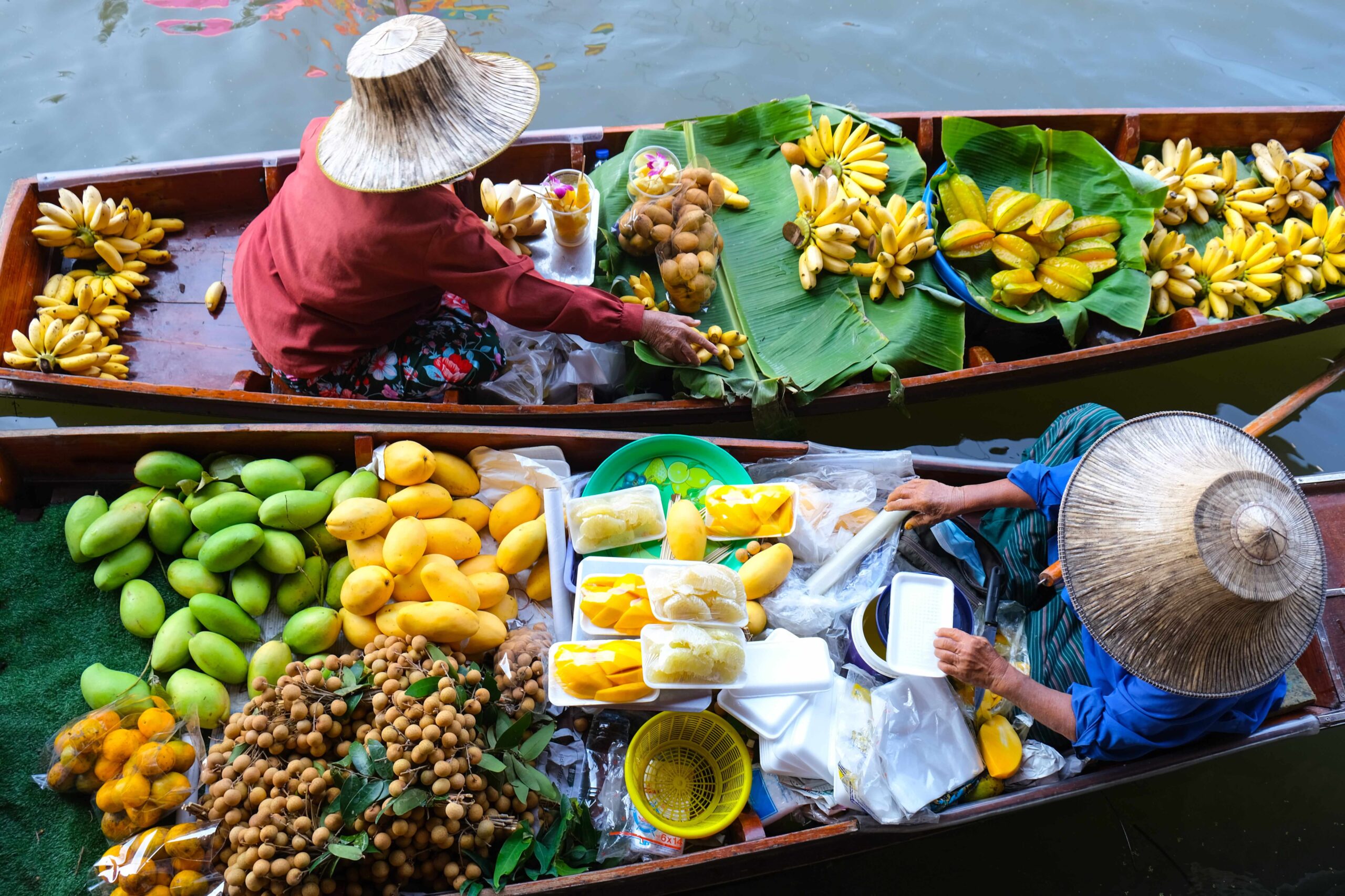 Aerial view of two wooden boats at a floating market with people selling fresh fruits such as bananas, mangoes, and other produce arranged on large green leaves.)