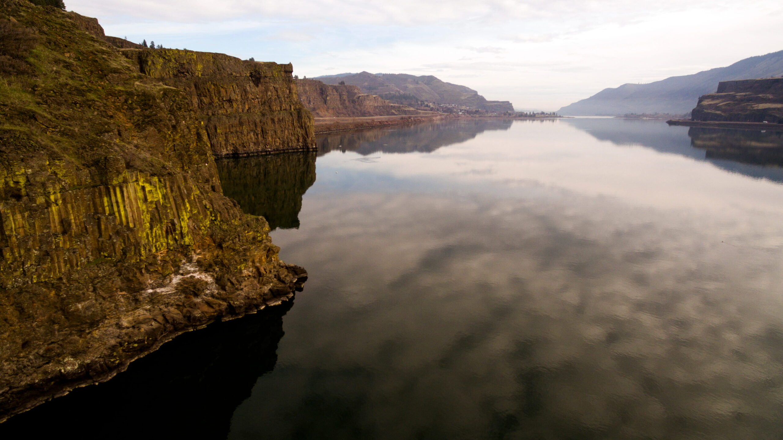 View of the Columbia River Gorge near Horsethief Butte, with basalt cliffs rising above calm, reflective water under a soft overcast sky.